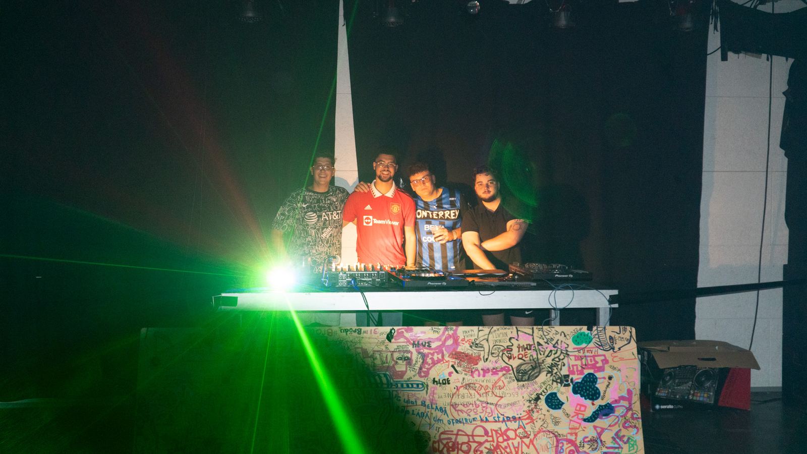Wide shot of a DJ booth in a dark room with a bright green spotlight flaring in front of the decks.